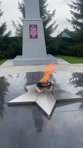 The video shows a memorial dedicated to the soldiers who fought in the Great Patriotic War, also known as World War II. The memorial consists of a grey obelisk with a red plaque on the front. The plaque contains text written in Russian, but it is not legible in the video. Below the obelisk is a black marble surface. In the center of the marble surface is a metal star-shaped structure with a small, circular opening in the center. Flames are burning in the center of the star. The flame is yellow and orange.  The background is composed of green grass and dark green evergreen trees. In the distance, a house is barely visible. The sky is overcast. The camera is stationary.