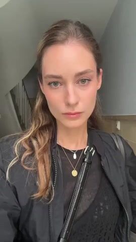 A woman stands in a stairwell. She wears a black jacket, a black sheer top, and a dark skirt with a pattern. She has two bags, one slung over her shoulder and the other held in front. The stairwell has a tiled floor and a railing of white balusters. She purses her lips and closes her eyes briefly.
