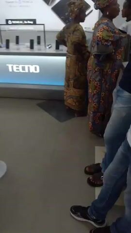 The video shows the interior of what appears to be a TECNO mobile phone store. A display counter with illuminated TECNO branding is prominent. Several mobile phones are displayed on the counter. A banner behind the counter advertises TECNO products like the Phantom V Fold2 and Camon 50 series. Several people are browsing and waiting in the store. Two women in brightly patterned dresses and headwraps stand near the counter. Men in casual attire are present, one in a white t-shirt looking towards the camera. A few employees in yellow shirts can be seen attending to customers. The overall environment is well-lit.