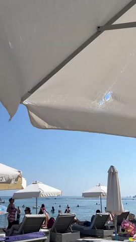 The video shows a beach scene on a sunny day. In the foreground, a large white umbrella covers a portion of the view. Underneath the umbrella, several empty lounge chairs are visible on the sand. Further back, the sea stretches out under a clear blue sky. People are seen swimming and relaxing in the water. There are other beach umbrellas and lounge chairs set up on the sand. The atmosphere is relaxed and leisurely, typical of a beach resort.
