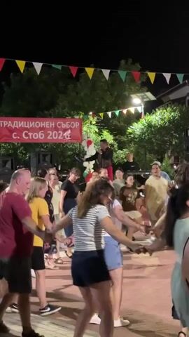 A crowd of people are dancing in a circle at night. There are colorful flags strung overhead. A banner in the background reads "ТРАДИЦИОНЕН СЪБОР С. Стоб 2022" (Traditional gathering of s. Stob 2022). A person is standing on a platform in the center of the dancing circle, possibly a performer or speaker. Several speakers are set up to the left of the platform. The ground appears to be paved. The lighting is artificial, coming from streetlights and possibly stage lighting. Some people are holding hands and dancing in a traditional circle dance. The atmosphere appears festive.