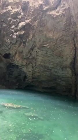 The video pans across the rough, textured rock walls of a cave. A pool of clear, turquoise water occupies the bottom of the frame, with scattered white objects visible beneath the surface. A mosaic icon is embedded in the cave wall, depicting a figure in blue and white robes surrounded by smaller figures. The lighting is dim, with sunlight filtering in from above. The camera movement is smooth and slow, creating a sense of exploration and reverence.