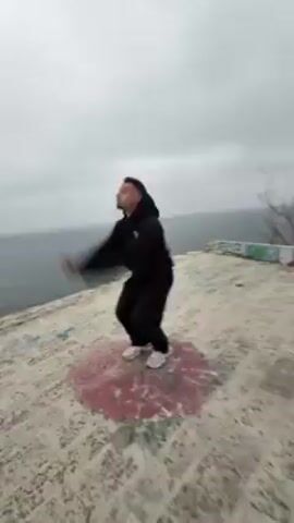 The video shows a person performing a backflip on a concrete surface next to the ocean. The person is wearing black pants, a black jacket, and white shoes. The surface they are on is covered in graffiti and has a pink circular design on it. The ocean is visible in the background, and the sky is overcast. After the flip, the camera pans to show the ocean and the rocky coastline below. The waves are crashing against the shore, and there are trees growing on the cliffside. There is more graffiti on the wall.