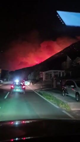 The video shows a nighttime scene from the perspective of someone in a vehicle. The vehicle is on a road that appears to be paved, with lane markings visible. In the distance, a hillside is engulfed in a large wildfire. The flames are a vivid red and orange, illuminating the smoke that billows into the night sky. The fire appears to be spreading across the slope, with visible points of light suggesting active burning. On the right side of the road, there is a building with a covered outdoor seating area. Several vehicles are visible on the road ahead of the viewer, with their headlights on. The overall impression is of a dramatic and potentially dangerous situation.