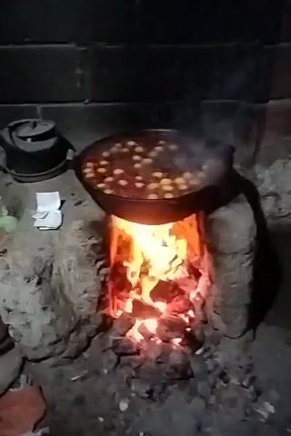 A black skillet filled with what appears to be meatballs in a red sauce is suspended over a wood-burning fire. The fire is contained within a rustic, stone-built stove, with glowing embers and bright flames visible. Wisps of steam rise from the cooking food, suggesting it is hot and actively being prepared. To the left of the skillet, a black teapot with a lid is placed on a surface, and next to it are two small white folded items, possibly napkins or paper. The background appears to be a dark, brick-like wall, and the overall lighting suggests a dimly lit environment, possibly evening or indoors. The scene is set outdoors, in a rustic, possibly rural location, given the primitive cooking method. The activity is clearly cooking. There are no people visible. The overall impression is one of traditional, home-style cooking.