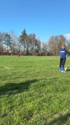 A soccer game is in progress on a green grass field, with a few players and a goal visible in the distance. A young boy in a blue soccer uniform with "ОК-СЕВЛИЕВО" on the back is standing on the sidelines next to a soccer ball, watching the game. Near him lies a small dog on a leash next to a bag and a water bottle. A woman in a red vest approaches and stands next to the boy, also watching the game. The sky is blue with some white clouds, and there are trees surrounding the field.