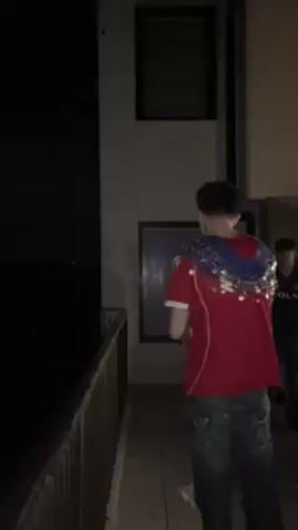A young man in a red and blue shirt and jeans is opening a bottle of champagne in front of a group of people. He is holding the bottle with both hands and shaking it vigorously, causing the champagne to spray out. The people behind him are watching and cheering. The event takes place on a patio or balcony with a metal railing, outside a building at night.