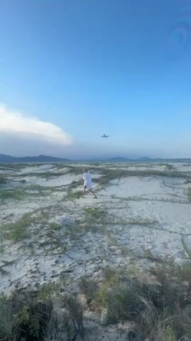 A person stands in a sandy, vegetated landscape under a bright blue sky. An airplane is visible in the sky above the person and the landscape. The landscape appears to be a coastal area with dunes and low-lying vegetation. The person is wearing a white garment. The airplane is in flight and appears to be descending. The sun is shining, casting a bright light on the scene.
