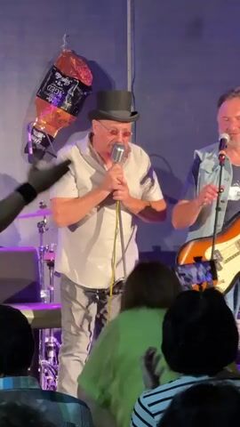 A man in a light grey short-sleeved shirt, light grey pants, round glasses, and a black top hat is standing center stage, holding a microphone up to his mouth. He is singing or speaking into the microphone. To his right, a man in a denim vest over a dark blue t-shirt, holding a guitar, is also facing forward and smiling. To the left of the man with the top hat, a person in a black t-shirt and jeans is partially visible, seated at a drum kit. In the foreground, a person wearing a bright green top faces away from the camera, with their back to the stage. Behind them, a seated audience is visible, some with curly hair. The stage has a plain blue backdrop and various musical equipment, including microphones, guitars, and drums. A large, gold-colored inflatable bottle, possibly champagne, is hanging from the ceiling to the left of the man in the top hat. The lighting is dim and appears to be stage lighting. The video pans slightly to the left and then back to the right.