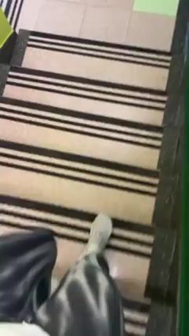 The video shows a person walking down a set of stairs. The stairs have a carpet runner with black stripes. The landing is tiled with a combination of beige, gray, and green tiles. The walls are painted green. At the bottom of the stairs, there's a large window that looks out onto a snowy street with people and cars. The person is wearing tie-dye pants. The camera is held by the person descending the stairs, providing a first-person perspective.