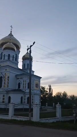 The video shows an external view of an Eastern Orthodox church at dusk. The church is painted light blue and has gold domes topped with crosses. The sky is a mix of gray and orange from the setting sun. A white picket fence surrounds the church. Power lines are visible crossing the sky. There is a road adjacent to the church. Some trees are visible in the background.