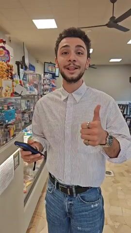 The video captures a young man demonstrating a contactless payment in a retail store, likely a convenience store or "tabaccheria" in Corsico, Italy.

The main subject is a young man with short dark hair, a beard, wearing a white striped button-up shirt, blue jeans, a black belt, and a watch. He has a ring on his left hand and an earring. At the beginning of the video, he smiles at the camera, gives a thumbs-up gesture, and holds up his smartphone, which displays a payment interface with a "Visa" logo and the text "Hold near reader."

He then approaches a counter where another person, likely a cashier with short dark hair and glasses, stands behind a cash register. The young man taps his smartphone onto a black, flat payment terminal on the counter. The phone screen briefly shows a processing animation before indicating a successful transaction.

The store's interior features shelves stocked with various products, including snacks, lighters (some branded "Clipper"), and other small items. On the counter, there's a sign in Italian that reads "Acquista QUI i tuoi codici" (Buy your codes HERE), advertising gift codes for services like Netflix, Xbox, Roblox, Twitch, Uber, Google Play, and Apple, indicating the store offers digital product sales. The overall lighting is bright, suggesting an indoor setting during the day.