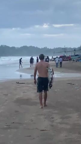 A man walks away from the camera carrying a surfboard on his right side. He is wearing dark shorts and is shirtless. The man is walking on a sandy beach toward the ocean in the background. Several other people are visible on the beach and near the water. There are boats parked on the beach in the distance. The sky is overcast and the lighting suggests it is either dawn or dusk. Trees and vegetation line the background of the beach.