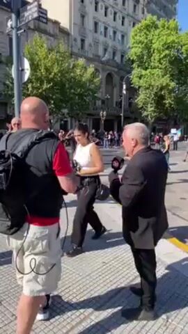 The video shows a crowd of people gathered in the street, with two men in the foreground who appear to be media reporters. The man on the left is wearing a red shirt, light-colored shorts, and a black backpack. He holds a camera and focuses it on the crowd. The man on the right is wearing a dark suit and holds a microphone with a logo on it, also directing his attention towards the crowd. The street is lined with buildings, some of which have visible signs. Trees are scattered along the street, adding some greenery to the urban environment. The crowd includes people of various ages and appearances, some standing still and others moving around. Debris litters the street, suggesting that some sort of event or activity may have taken place recently. The overall atmosphere seems active, with the crowd and the reporters all engaged in observing or documenting the scene.