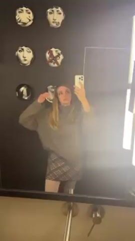 A woman with blonde hair and a gray turtleneck sweater is taking a selfie in a mirror. She's also wearing a plaid skirt and light-colored tights. The background includes a dark wall decorated with small, round plates featuring facial images. She poses, touching her hair and making a kissing face toward the camera. Her white smartphone is visible in her hand, and a ring is on her finger. The setting appears to be a bathroom or restroom, with a glimpse of a sink and fixtures at the bottom of the frame.