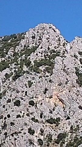 The video shows a rocky mountain peak against a clear blue sky. The mountain is covered in sparse green vegetation.
