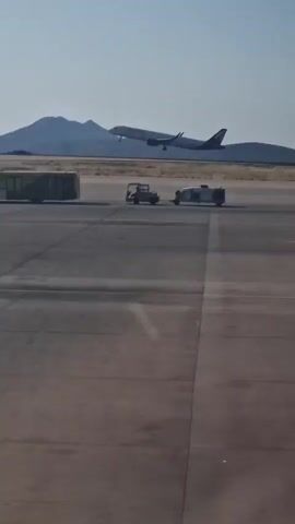 An airplane is ascending into the sky against a clear blue backdrop, with hills visible in the distance. In the foreground, a Hertz shuttle bus is parked on an asphalt surface, likely an airport tarmac. A small utility vehicle is also present nearby. The scene captures a moment of air travel, with the plane in mid-flight and ground transportation visible.