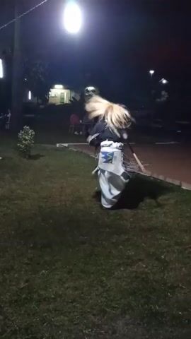 At night, in an outdoor area illuminated by a bright overhead light, a figure in a white and black outfit with a straw-like headdress dances rhythmically. The individual twirls and sways, holding long objects that resemble poles. The scene takes place on a grassy lawn adjacent to a reddish-brown pathway next to a swimming pool. Tables with seated individuals are visible in the background, creating a public or event-like atmosphere. The light source casts strong shadows, accentuating the dance against the dark background.