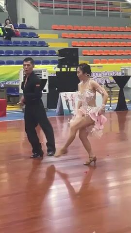 A man and a woman are dancing ballroom dance in a stadium. The man is wearing a black shirt and pants, and the woman is wearing a pink dress. They are on a wooden floor, and there are bleachers in the background. The woman is spinning, and her dress is flying out. The man is looking at her and smiling. There are some banners and speakers in the background. The lighting is bright, and it appears to be daytime.
