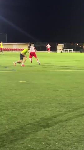 The video shows a group of young males on a bright green, artificial turf soccer field at night. The field is illuminated by overhead lights, creating strong shadows. There are several players in red jerseys and white shorts, and at least one player in a yellow-green jersey and dark shorts. They appear to be engaged in a soccer practice or game, as players are running, kicking a ball, and positioned on the field. A soccer goal is visible in the background, as is a line of empty bleachers with a fence behind them. The sky is dark, indicating it is nighttime. The camera is handheld and slightly shaky, following the action on the field.