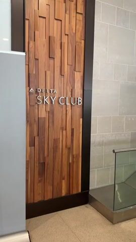 The video shows the entrance to a Delta Sky Club lounge. The entrance features a wooden wall with the words "DELTA SKY CLUB" in silver lettering. To the right of the entrance, there is a glass wall with a blue geometric pattern and the Delta Sky Club logo. In the foreground, there is a grey rolled-up banner on the left and a glass railing on the right. The video pans across the entrance, showing different angles of the signage and the surrounding area.