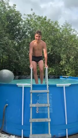 A young boy in black swimming trunks climbs the ladder to an above-ground swimming pool. He stands at the top of the ladder, holding onto the rails, and extends his arms outwards. He then jumps into the pool, creating a large splash. After the splash subsides, the boy's head emerges from the water, and he covers his face with his hands. The pool is blue, and surrounded by green trees and foliage.