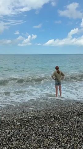 A person stands on a pebble beach facing the ocean with waves coming in. The person is wearing a green t-shirt and denim shorts. They have their hands on their hips and their feet are in the water. The sky is blue with scattered clouds.