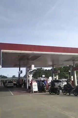 The video shows a gas station with several cars and motorcycles lined up to refuel. The sky is clear and the sun is shining. There are trees and buildings in the background. A person is standing next to a sign that says "BBM". The gas station appears to be busy.