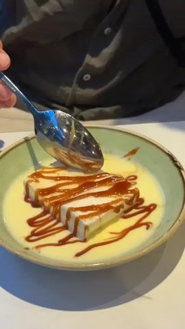 A close-up shot shows a spoon scooping a piece of white dessert drizzled with caramel sauce from a bowl. The dessert is rectangular and sits in a creamy yellow sauce. The bowl has a green rim. The background is blurred, but a person wearing a dark shirt is visible.