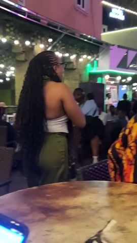 A medium shot captures a woman with long, dark braided hair pulled into a ponytail, wearing a white halter top and olive-green pants, standing with her back to the camera. She is in an outdoor setting, possibly a patio or rooftop bar, with string lights overhead and lush greenery visible in the background. Several other people are present, some blurred in the foreground and background, suggesting a social gathering or event. A man in a yellow jersey with the number 23 is visible further back. The overall atmosphere appears lively and casual.