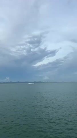 The video shows a body of water under a cloudy sky. There are two jet skis in the distance, creating wakes on the water. In the far distance, there is land, which appears to be hilly. The water is a deep green color, and the waves are small. The clouds are a mix of white and gray, with some darker clouds in the distance that suggest the possibility of rain. The overall mood of the video is calm and serene.