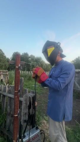 A person wearing a welding mask, gloves, and a blue long-sleeved shirt is welding a metal post. They are holding a welding torch and sparks are flying from the point of contact with the post. A welding machine is visible at the base of the post, with wires connected to the torch. The scene appears to be outdoors in a grassy area with a wooden fence in the background and trees in the distance. The sky is clear and blue. The video is a short clip of the welding process.