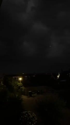 A very dark video shows a dimly lit residential area at night. The sky is cloudy and dark, with faint light illuminating some of the clouds, suggesting a storm or overcast conditions. In the foreground, there are dark silhouettes of trees and bushes, with some white flowers visible at the bottom left. A path leads into the scene, illuminated by a single streetlamp. In the background, the dark outlines of houses with a few lit windows can be seen. The overall atmosphere is dark and quiet.