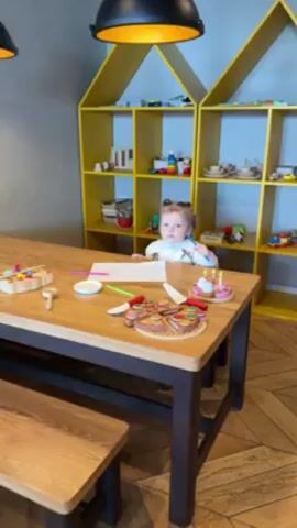 The video shows a young child sitting at a wooden table with a wooden bench. The child has blonde hair pulled into pigtails and is wearing a cream-colored sweater. The child is drawing on a piece of white paper with a blue marker. On the table are various wooden toys, including a pizza and a cake. Behind the table is a yellow bookshelf shaped like houses, filled with various toys and objects. The lighting in the room is bright.