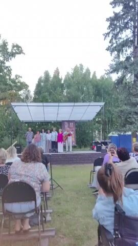 A group of people are standing on a raised outdoor stage under a white canopy. The group consists of five individuals, three women and two men, all dressed casually. One of the men is holding a guitar and appears to be playing it. The stage is set in a grassy area surrounded by trees, suggesting an outdoor performance or event. In the foreground, an audience is seated on chairs, facing the stage. Some members of the audience are visible from behind, and one person in the front row is holding up a phone, possibly to record the event. The lighting suggests it is either late afternoon or early evening.