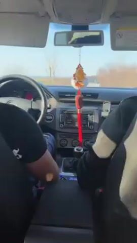 The video shows a car's interior from the perspective of the driver's seat. The car, a Volkswagen Passat with a manual transmission, is traveling down a two-lane road. The driver is visible from the shoulders down. They are wearing a black t-shirt and jeans. A passenger's leg, clad in black pants, is visible next to the center console. The dashboard features an integrated car stereo, air conditioning vents, and an emergency flasher button. A decorative tassel hangs from the rearview mirror. On the road ahead, a convoy of cars is visible in the distance. The scenery outside the car includes grassy fields and trees. The weather appears sunny. Music with a heavy bassline plays throughout the video.
