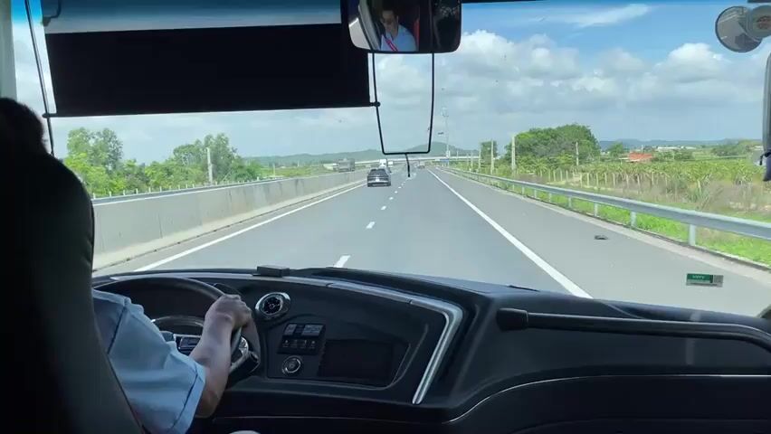 The video provides a first-person perspective from inside a bus, looking forward through the windshield. The driver's hands are visible on the steering wheel, suggesting they are actively driving. The bus is on a highway, with clear lane markings and guardrails visible. Other vehicles, including trucks and cars, are seen in the distance on the highway. The surrounding environment is rural, with green fields, trees, and some vegetation visible on either side of the road. The sky is blue with scattered clouds. A rear-view mirror reflects an image of a person wearing a seatbelt. The dashboard of the bus is also visible, showing various controls and a screen. The overall scene depicts travel on a highway in daylight.