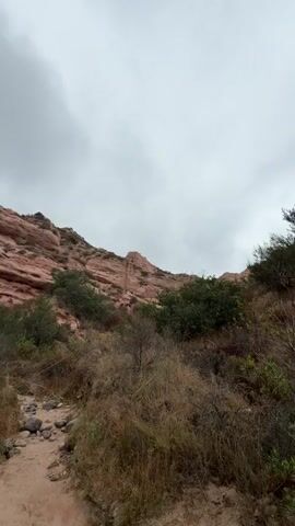 This media file captures a low-angle, wide shot of a hiking trail winding through a natural landscape under an overcast sky. The scene is dominated by the earthy tones of the terrain and the warm hues of the reddish-pink rock formations that rise prominently on the left and in the background.

The trail itself is a sandy path, dotted with small stones and pebbles, leading upwards and away from the viewer. It is bordered on either side by dry, straw-colored grasses and sparse green bushes, indicating a somewhat arid environment. The vegetation appears dry and weathered, typical of a region that might experience periods of drought.

The prominent rock formations are layered and striated, suggesting geological history and erosion. They form a rugged cliff face that reaches towards the sky. Patches of darker green foliage are visible clinging to the lower slopes of these rocks and scattered throughout the mid-ground, providing a contrast to the dry grasses and rock.

The sky is filled with heavy, grey clouds, suggesting a day that is either cloudy, preparing for rain, or has recently experienced it. There are no people or man-made objects visible in the frame, emphasizing the natural and somewhat remote character of the location. The overall mood is one of natural beauty and the quiet solitude of a hike in a rugged, outdoor environment. The time of day is indeterminate due to the overcast conditions, but the lighting suggests it is daytime. The location cues, such as the red rock formations and arid vegetation, are suggestive of the American Southwest, and the context provided places it in Lake Forest, United States of America.