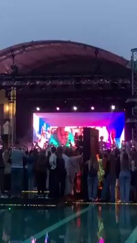 A crowd of people is gathered to watch a live performance on an outdoor stage. The stage is covered by a roof supported by metal scaffolding, and illuminated by bright spotlights and a large video screen displaying images of the performers. The audience, mostly young, is facing the stage, holding up phones to record the performance. A body of water separates the audience from the stage, reflecting the lights and activity. The scene is set against a dusky sky.