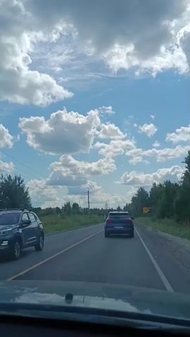 The video shows a view from inside a vehicle traveling on a paved road. A dark-colored sedan is visible ahead in the same lane. The road is flanked by grassy shoulders and trees. Power lines and poles are present on the left side of the road. The sky is blue with scattered white, fluffy clouds, and the sun is visible through a gap in the clouds. A yellow sign is partially visible on the right side of the road. The camera appears to be positioned on the dashboard or windshield of the vehicle.