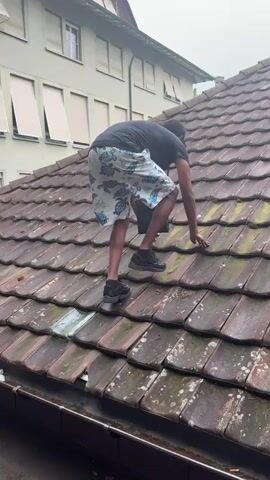 A man climbs on a red-tiled roof, reaching down to touch a tile. He is wearing a black shirt, shorts with a turtle pattern, and black shoes. He appears to be carrying a small black box. Another man stands in front of a building, partially blocking the view of the man on the roof. The building is numbered "1" and has white walls. The weather appears to be overcast. The second man briefly raises his hand towards the camera, obscuring the view.