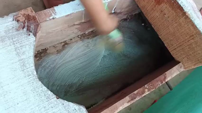 A close-up shot shows a person's hand holding a paintbrush with a green handle and white bristles. The paintbrush is applying a white substance, possibly paint or plaster, to the inside of a wooden structure. The wooden structure appears to be old and weathered, with visible grain and some rough edges. The interior surface being painted is rough and uneven. The surrounding environment is out of focus but appears to be outdoors with a concrete or dirt ground. The lighting suggests daytime.