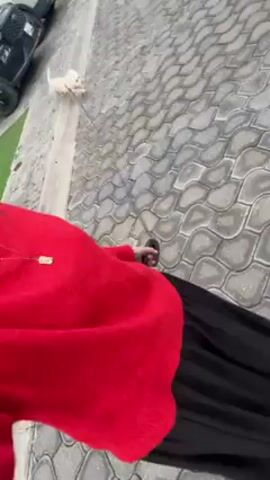 The video shows a person walking a small, white dog on a leash. The person is wearing a red sweater and a long black skirt, and sandals. They are walking on a paved surface made of interlocking blocks. A car is partially visible on the left side of the frame. The dog is running and pulling on the leash.