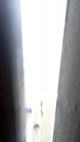 The video shows a beach from a balcony, moving from inside a building to reveal the scene. The beach has dark sand and several large rocks scattered along the shoreline and extending into the water. Small waves are visible lapping against the sand and rocks. In the distance, the ocean stretches to the horizon under a hazy sky. A lone person is walking along the beach. The balcony has white walls with blue-and-white tiled trim.