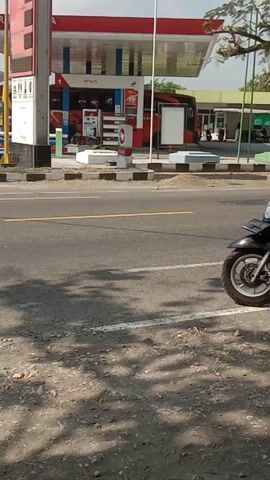 A gas station is visible on the left side of the road. A motorcycle is parked on the right side of the road. A truck is parked next to the motorcycle, facing away from the viewer. The truck is green and yellow and has the number TK102 on it. There is a person in the driver's seat of the truck. The road is asphalt and has a yellow line in the middle. There are trees and shadows on the right side of the road.