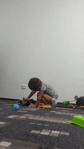 A young child is playing on the floor in front of a white wall. The child is wearing a t-shirt and a diaper. The child is crouching and appears to be interacting with toys on the floor. There are several toys scattered around the child, including a toy car, a xylophone, and some building blocks. The floor is carpeted.