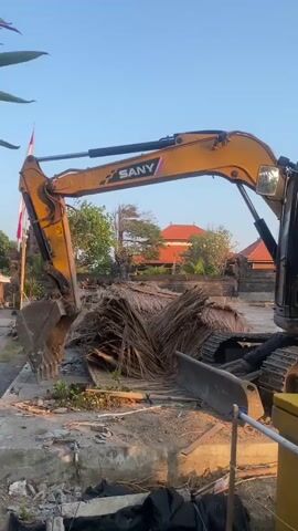 A yellow SANY SY75C excavator is parked on a construction site. The excavator is facing towards the right side of the frame. The boom of the excavator is raised and extended. A pile of dried palm fronds is located to the left of the excavator. In the background, there is a building with a red tiled roof and trees. The sky is clear and blue. The lighting suggests it is daytime, likely early morning or late afternoon due to the warm light.