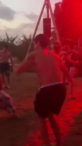 The video shows several people dancing on a beach, illuminated by red and purple lights. The focus is on a shirtless man in black shorts who is energetically jumping up and down, moving his arms while he dances. The surrounding people are also dancing, though their movements are less visible. The beach appears to be sandy, and there is some sort of structure visible in the background, made of wood. The sky in the background suggests it is dusk or early evening.