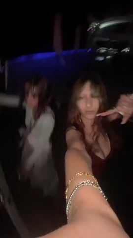 Two young women are seen in the foreground, seemingly on a moving platform, possibly a boat. The woman in the foreground on the right, wearing a dark red dress, is holding up her left arm as if taking a selfie. She has long, dark, wavy hair and is smiling. A beaded bracelet is visible on her wrist. Behind her and to the left, another young woman, wearing a white top and a light-colored skirt, is looking at the camera and smiling, with her right arm raised. The background is dark with blurred blue and white lights, suggesting a nighttime setting, possibly at a party or event. The perspective shifts to reveal a wide shot of a crowded boat at night. Many people are seated at tables on the deck, illuminated by yellow and blue lights. A large screen is visible at the front of the boat, showing a bright white light. The boat appears to be moving through water under a dark sky with distant lights visible on the horizon.