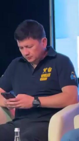 The video shows a man sitting in a chair and looking at his phone. He wears a black polo shirt with yellow text "MYKOLAIV REGION" and a trident symbol. He also wears a black smartwatch. He sits on a white armchair on a stage. Behind him, there is a large screen displaying an abstract pattern. A water bottle sits on the side table.